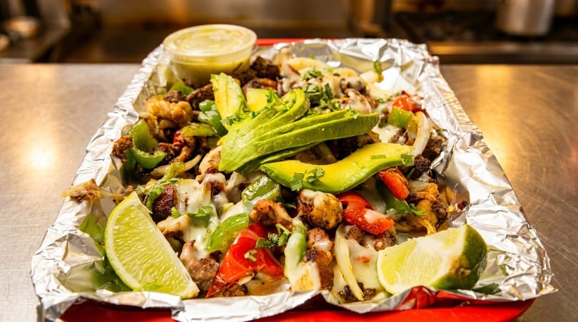 Foil-lined tray of fajita-style meat, peppers, onions, melted cheese, avocado, and lime wedges.